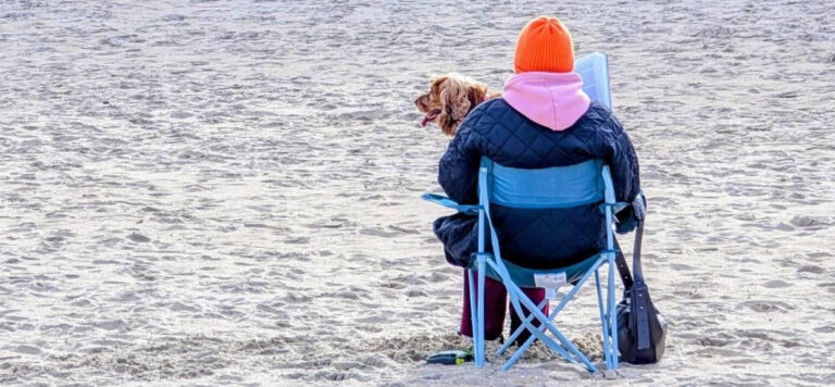 Kobieta siedząca na stołeczku na plaży z psem na kolanach wpatruje się w dal. Jak dobrze wyjść z mediów społecznościowych?