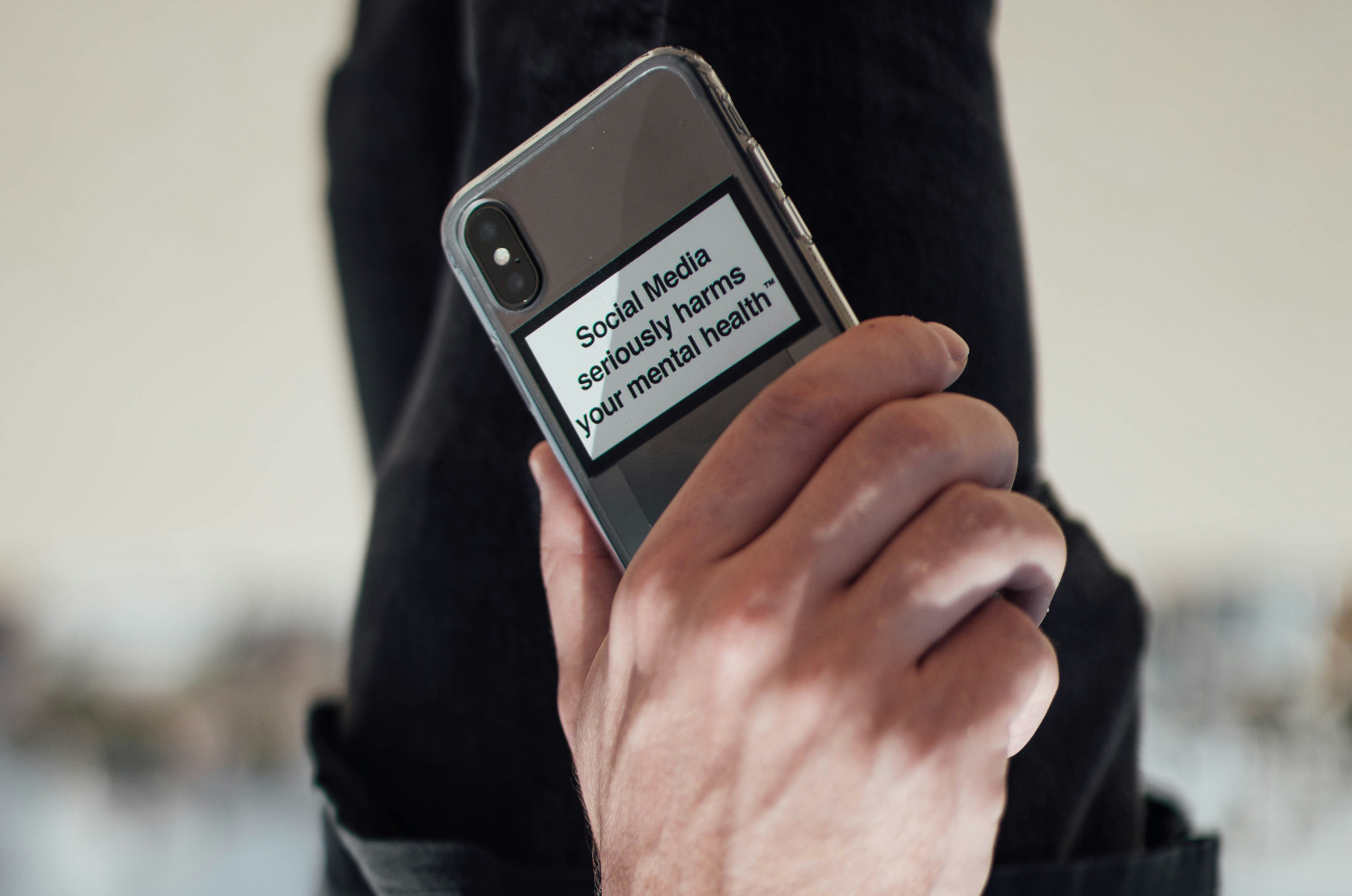 Ręka mężczyzny trzymająca telefon. Na tyle telefonu znajduje się napis. Media społecznościowe poważnie szkodzą zdrowiu psychicznemu.